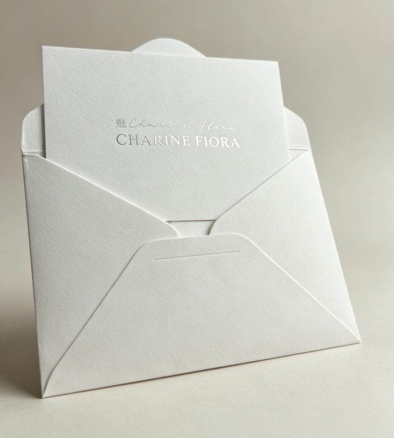 Silver foil embossed custom brand greeting card placed on textured white envelope, subject oriented left, premium thick cotton paper stationery for CHARINE FIORA brand packaging, minimalist dark background luxury branding accessory