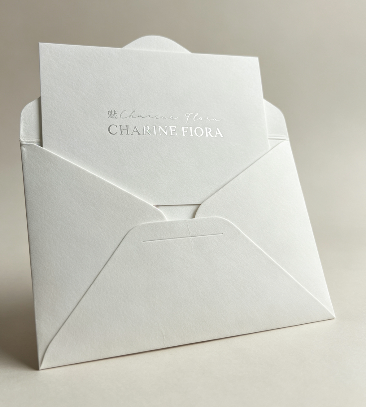 Silver foil embossed custom brand greeting card placed on textured white envelope, subject oriented left, premium thick cotton paper stationery for CHARINE FIORA brand packaging, minimalist dark background luxury branding accessory